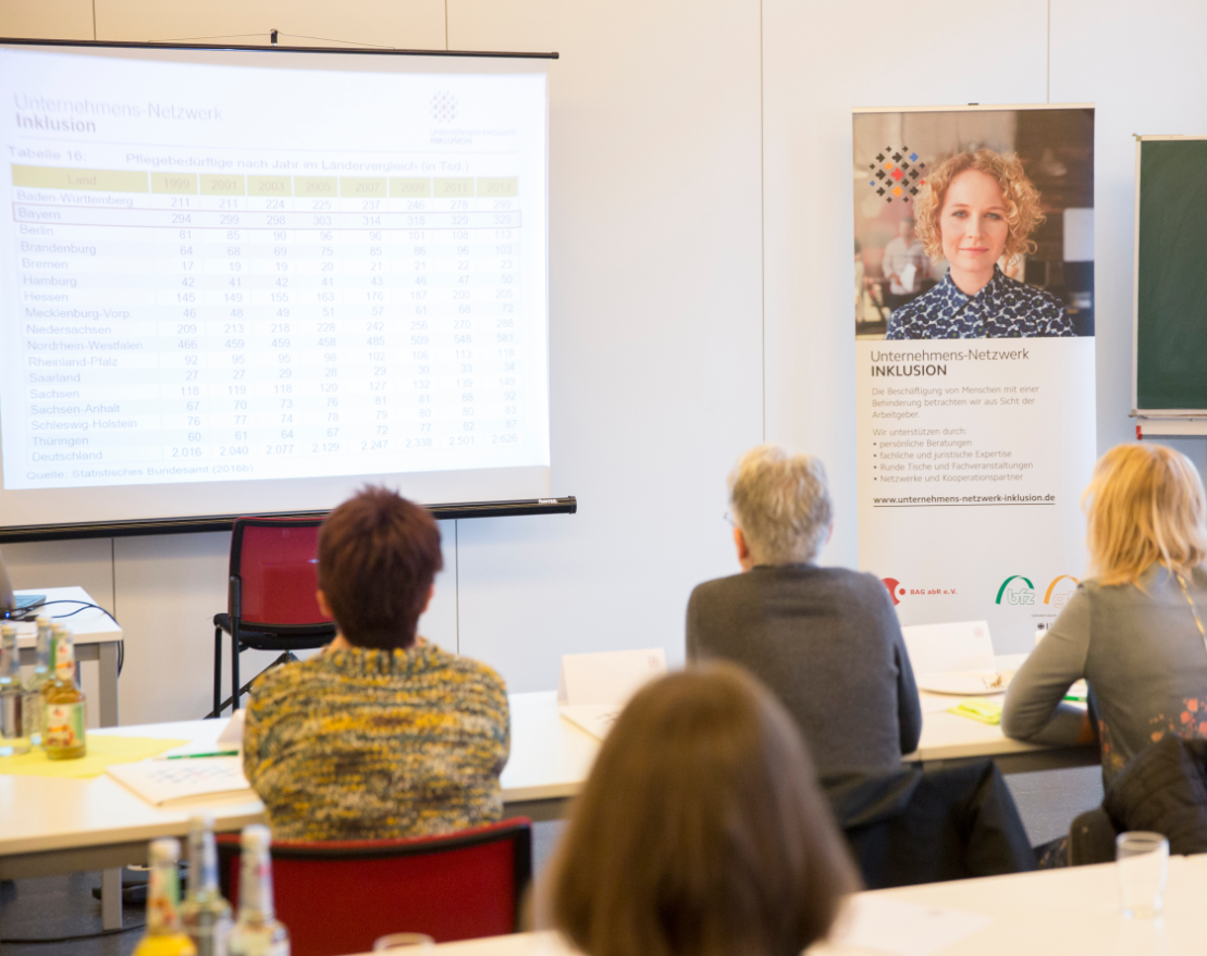 Menschen sitzen bei Veranstaltung mit Blick nach vorne auf Präsentation.